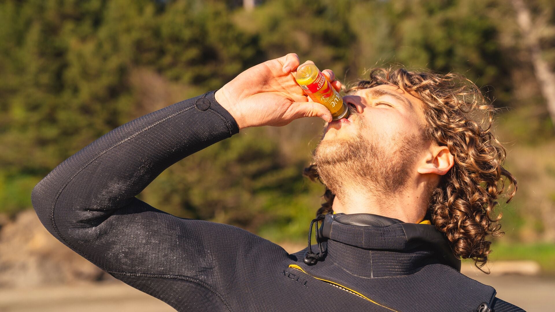 A surfer in a wetsuit drinking a Vive Organic Immunity Boost Original shot at the beach