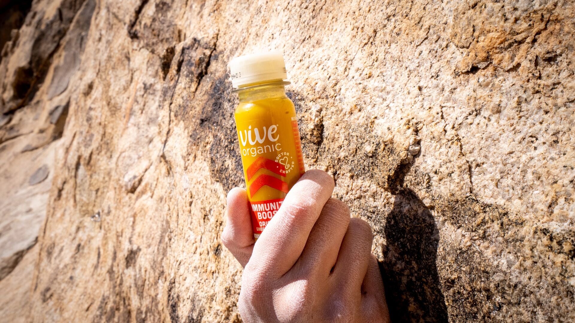 A hand gripping a sunlit rock face while holding a Vive Organic Immunity Boost Original shot