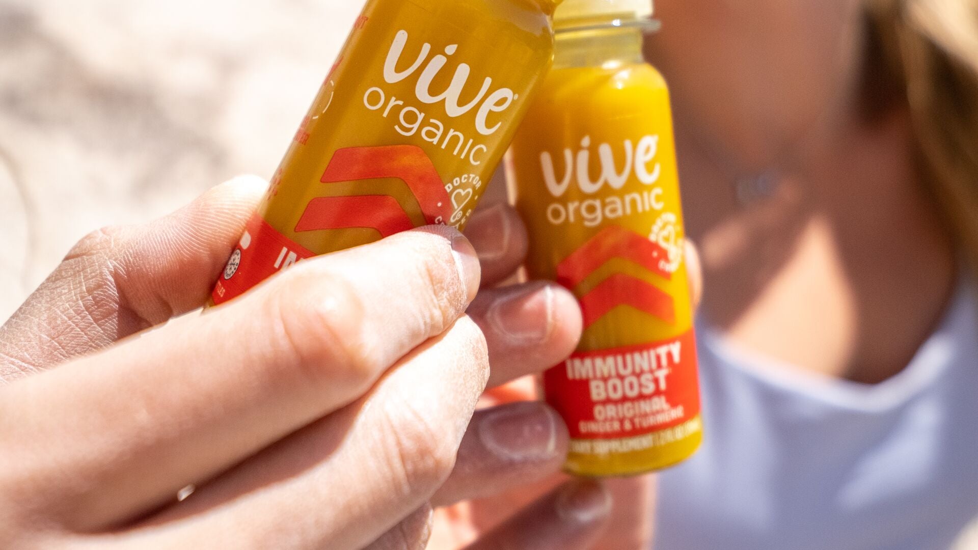 Two Vive Organic Immunity Boost Original shots held in the sun on a sandy beach