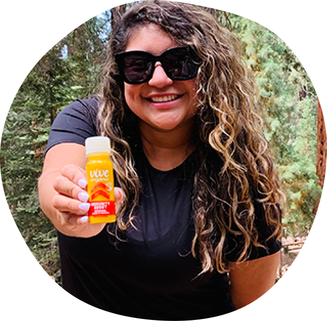 a woman smiling with sunglasses and long curly brown hair holding a vive organic immunity boost original shot in her hand.