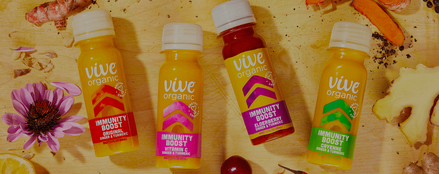 Image of Vive Organic Immunity Boost Shots collection laying flat on a cutting board surrounded by fresh echinacea flower, ginger, turmeric, acerola cherry, lemon and pepper.